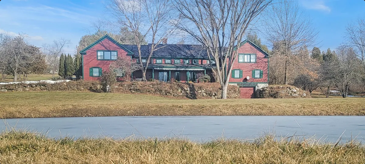 A classic view of the farmhouse with its red facade and green shutters, nestled beside a tranquil frozen pond and surrounded by bare trees. The setting is peaceful, hinting at seasonal changes and opportunities for outdoor gatherings or quiet moments of inspiration.