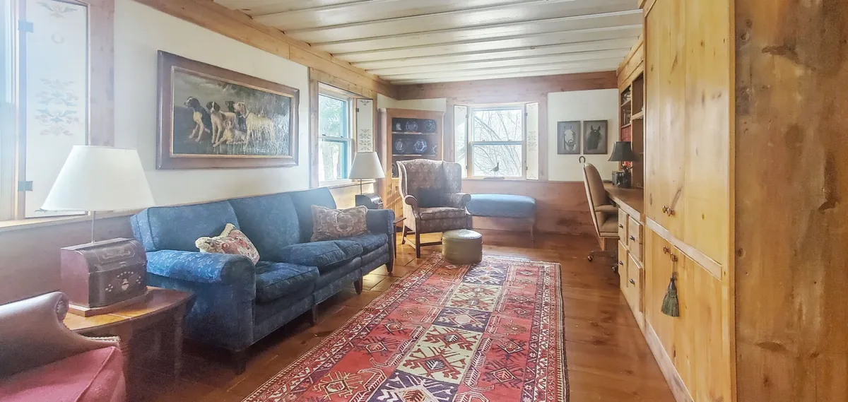 A welcoming room anchored by a comfortable blue sofa, a vibrant area rug, floor-to-ceiling wooden bookshelves stocked with books, and large windows letting in abundant natural light. This space is ideal for reading, relaxing, or working, and the windows suggest serene views beyond, aligning with the description of inspiring countryside vistas.