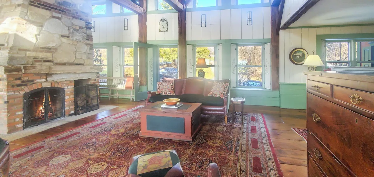 This image features a cozy living room with a fireplace, comfortable sofa, coffee table, a welcoming rug, and windows that offer a view of the outdoors. The ample natural light and the inviting setting suggest a tranquil environment perfect for relaxation or focused work.