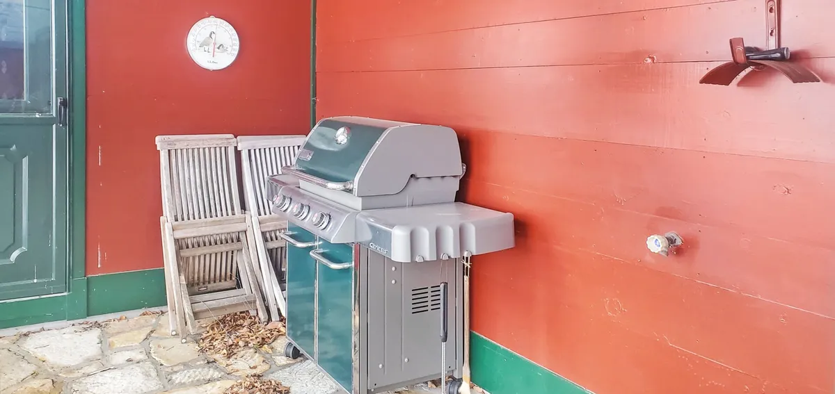 An inviting outdoor patio area with a grill, wooden chairs, and a striking red wall, set up for barbecuing, relaxing, and entertaining. The space is casual and welcoming, perfect for summer barbecues, evening hangouts, or enjoying a meal outside with a view of the landscape.