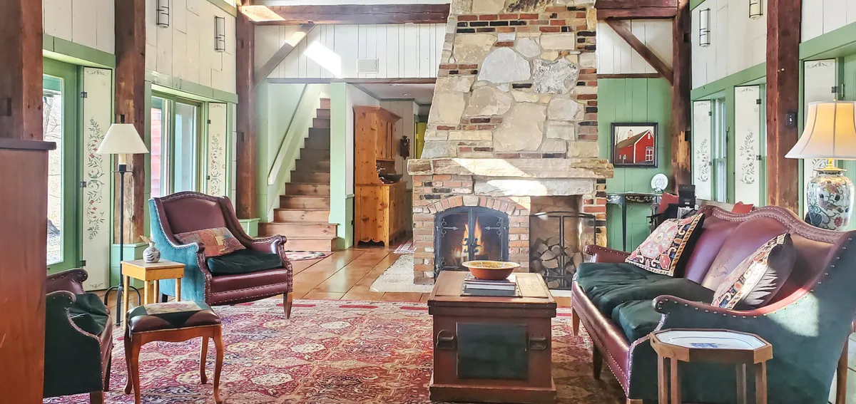 A warm, inviting great room featuring a dramatic floor-to-ceiling stone fireplace, comfortable seating, exposed vintage barn beams, and radiant pine wood flooring. Sunlight streams through ample windows, highlighting the rustic charm and providing a welcoming ambiance for gatherings and relaxation.