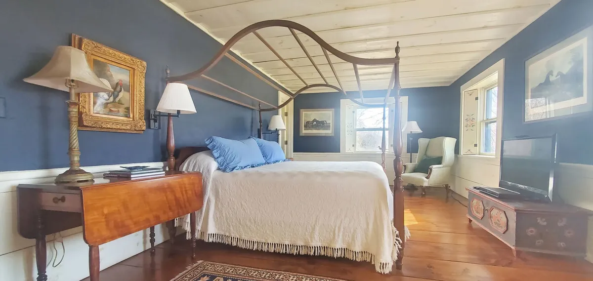 A tranquil bedroom with a four-poster bed accented by blue pillows, a wooden side table, a decorative lamp, framed artwork, and a comfortable armchair by the window. The space is bathed in natural light, suggesting a peaceful retreat for rest and relaxation.