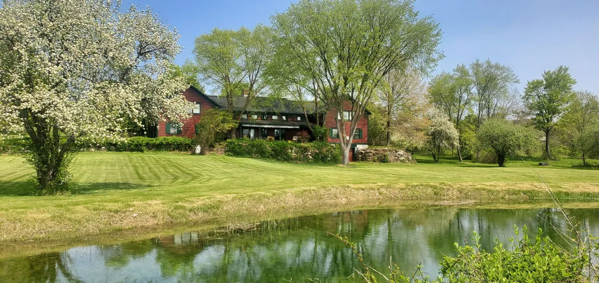 The image shows the charming Old Orchard Home nestled among blossoming apple trees, with a tranquil private pond in the foreground, lush green meadows stretching beyond, and vibrant flowers framing the scene. The spacious house is set against a backdrop of Vermont countryside, evoking a sense of peaceful seclusion and natural beauty.