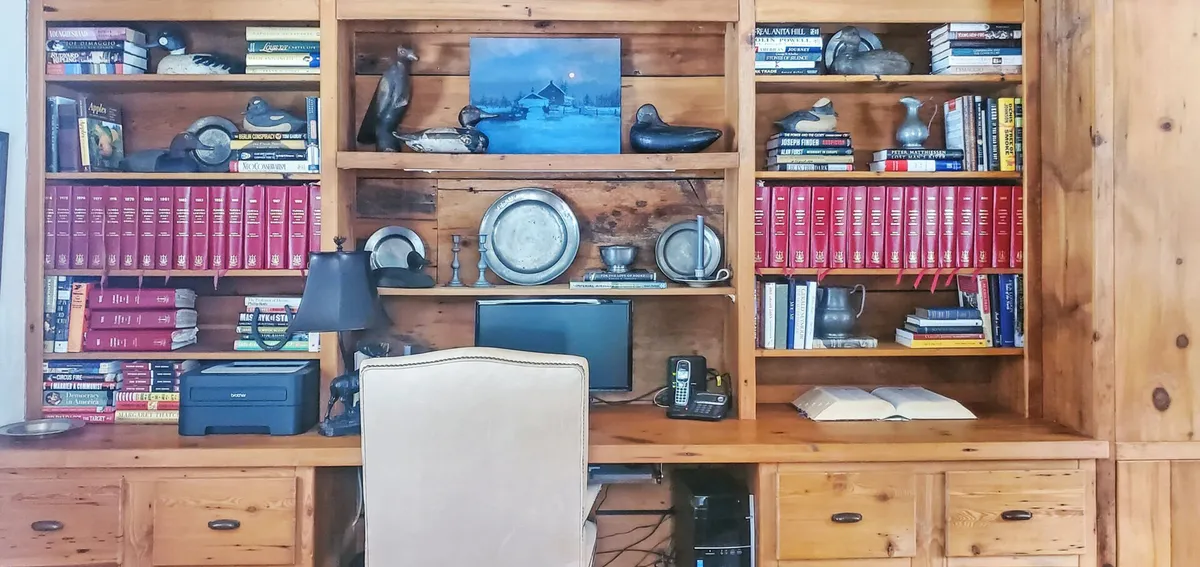 A cozy library/den featuring bookshelves filled with books, decorative duck figurines, framed artwork, and a dedicated desk area with a computer and lamp. The room is warmly lit and the wood elements give it a rustic, inviting feel. This image foregrounds both the intellectual and creative atmosphere while emphasizing the dedicated work-from-home setup.