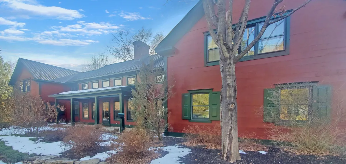 This image showcases the red exterior of the Old Orchard Home, accented by green shutters and surrounded by a landscaped garden. The scene evokes a welcoming and tranquil atmosphere, with the porch inviting moments of relaxation and the grounds suggesting gatherings or quiet contemplation.