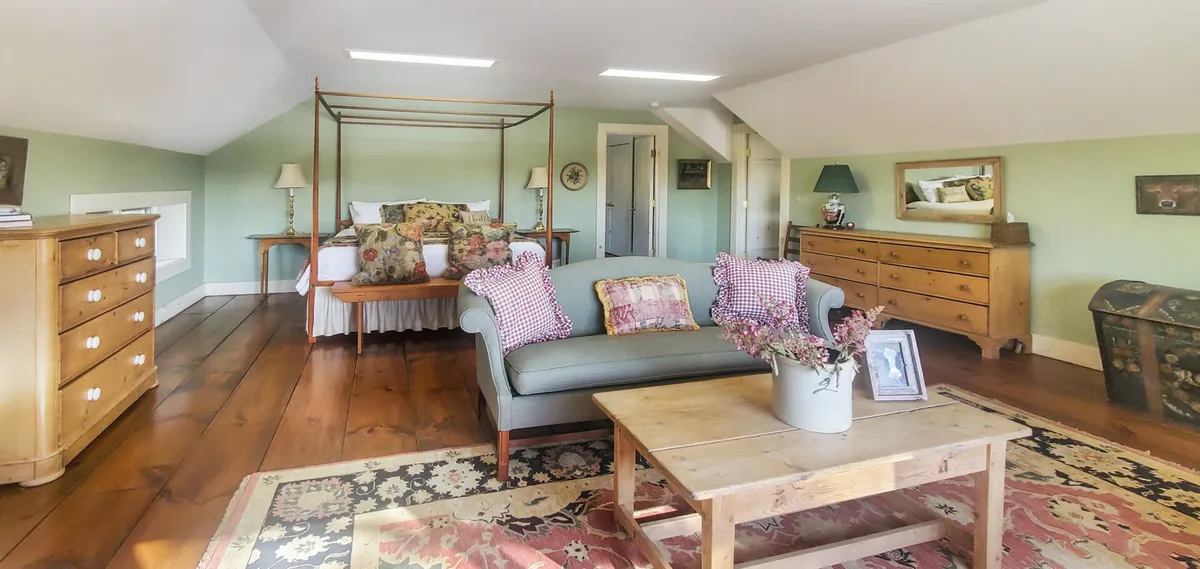 A light-filled bedroom featuring a canopy bed, a comfortable sofa, wooden dressers, a soft rug, and a vase with fresh flowers. Sunlight pours through the windows, creating a warm and inviting atmosphere that feels both restful and spacious.