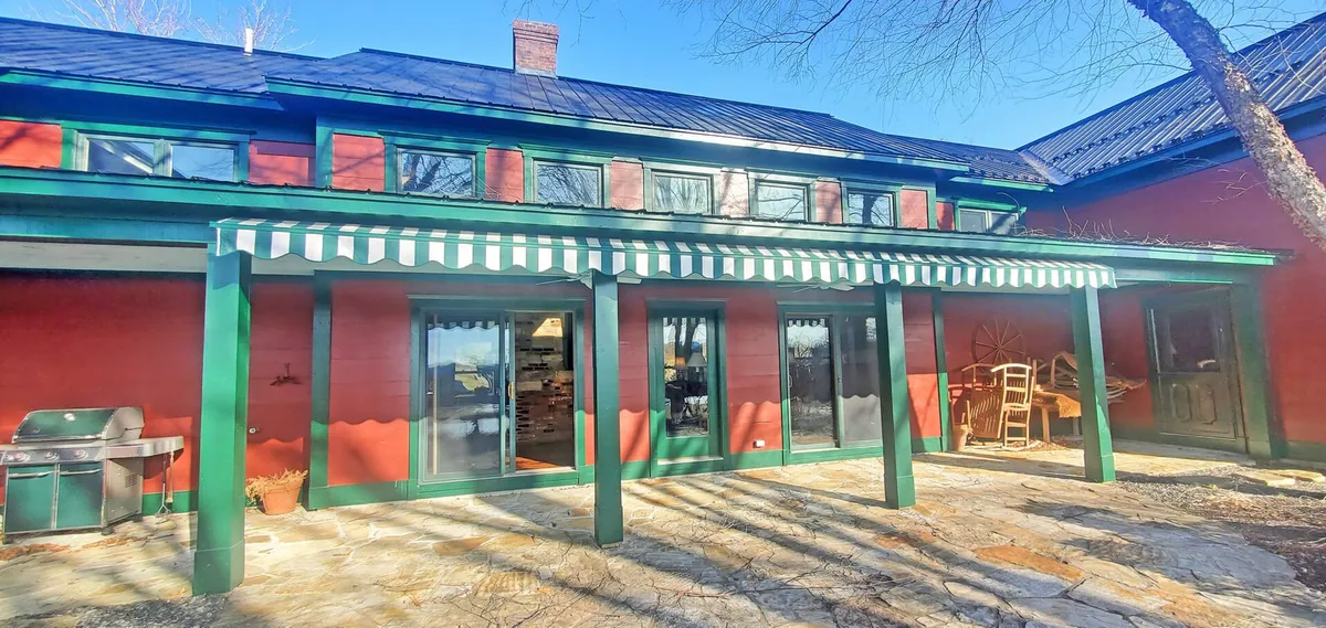This image showcases the estate's stone patio with large glass windows and a green striped awning, seamlessly connecting the indoor and outdoor spaces. The inviting patio setting hints at opportunities for alfresco dining, relaxing with a morning coffee, or enjoying sunset gatherings. The view through the glass doors highlights the natural flow between the home’s interior and exterior.