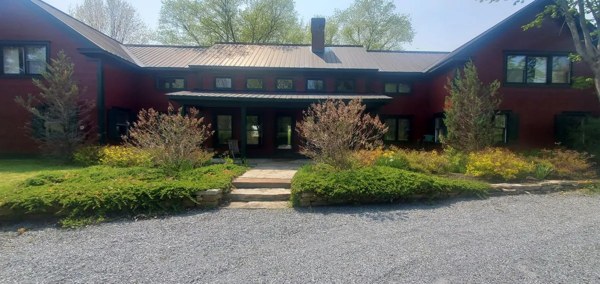 The image features the red exterior of the house, a manicured garden, and a welcoming stone pathway leading to the entrance. It captures the charm and hospitality of the estate, while also hinting at outdoor gatherings, garden exploration, and the experience of arriving at the property.