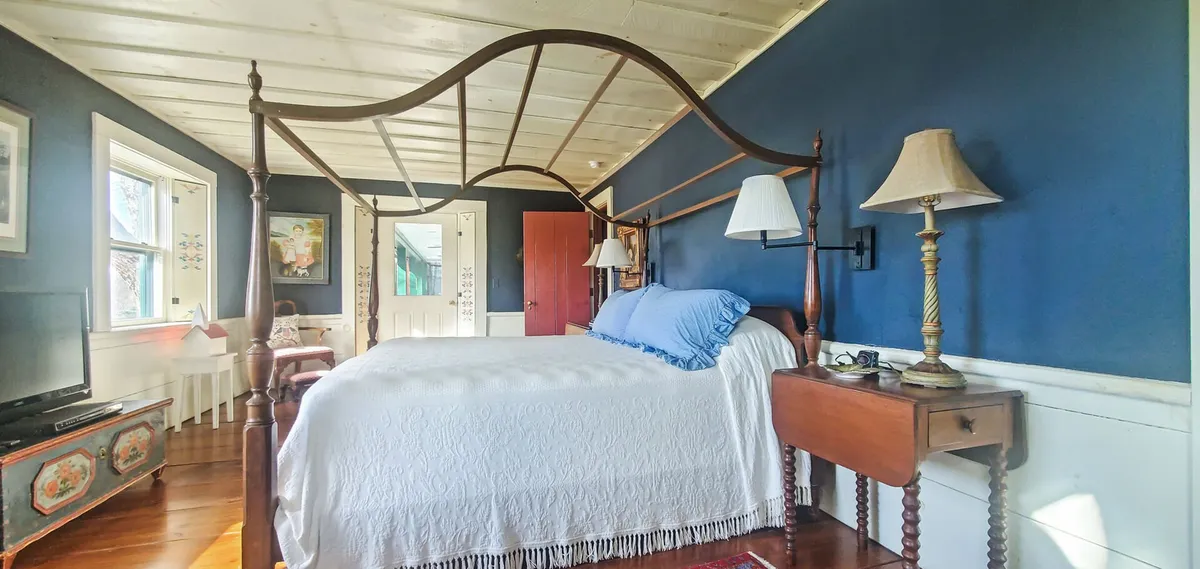 A tranquil bedroom with a four-poster bed, soft blue wall, wooden furniture, and gentle lamp lighting creates a calm, quiet atmosphere ideal for relaxation or focused thought. Natural light filters through the window, suggesting a bright yet serene ambiance.