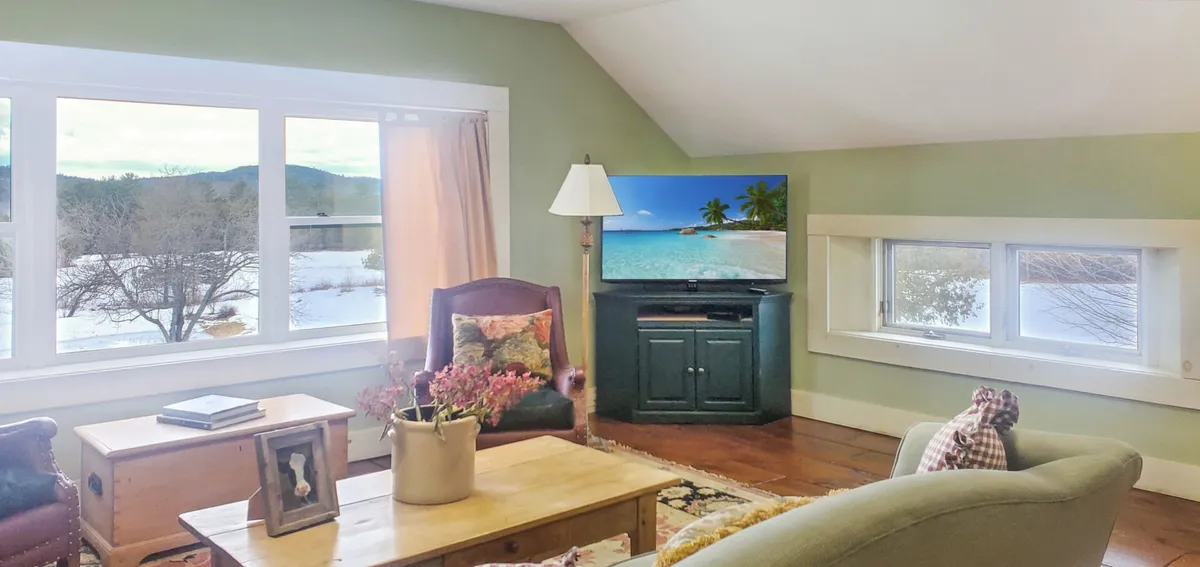 A cozy upstairs den with large windows that frame a mountain view, a comfortable seating area, and a television displaying a beach scene. The room is bright, airy, and perfectly suited for movie nights or quiet relaxation while taking in scenic vistas.