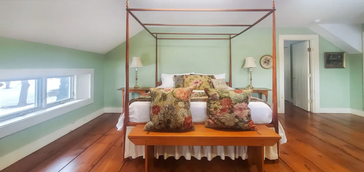 This serene bedroom features a canopy bed with decorative pillows, a wooden bench, large windows letting in natural light, and cozy touches inviting relaxation or meditation. The expansive windows frame views of the natural landscape outside.