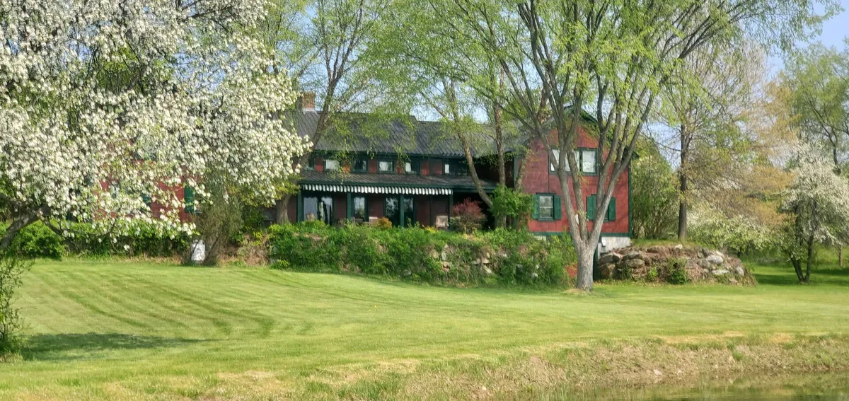 This image presents the estate's striking red house set against a backdrop of flowering trees and manicured green lawn. The tranquil exterior scene suggests spaces for children and pets to play, while the inviting porch hints at family gatherings or quiet moments overlooking the property.