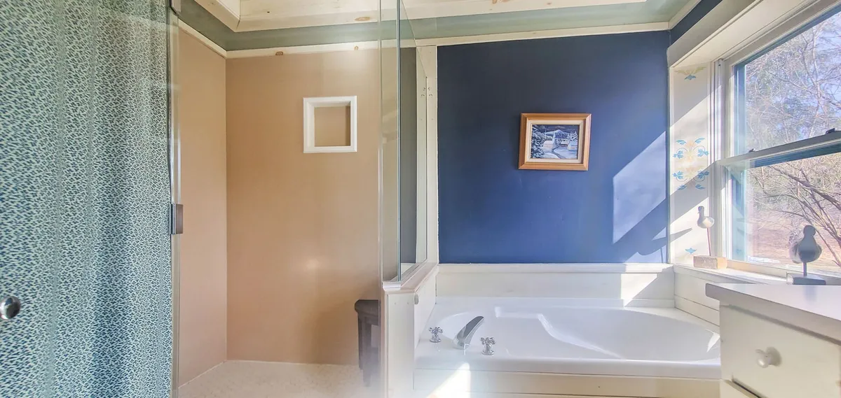 A serene bathroom featuring a spacious shower with a glass panel, a relaxing bathtub, framed artwork, and a window allowing in natural light. The scene is tranquil, evoking spa-like comfort and privacy.